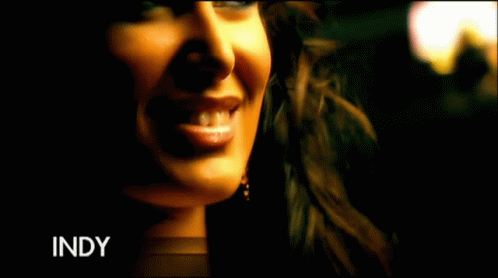A Woman In A Gold Dress Is Standing In A Room With The Word Indy On The Bottom Left GIF