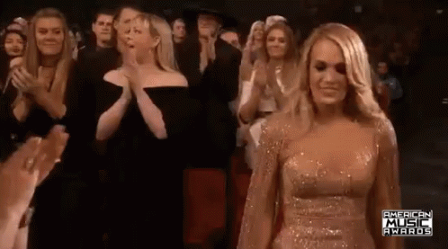 A Woman In A Gold Dress Is Standing In Front Of A Crowd Of People Clapping . GIF