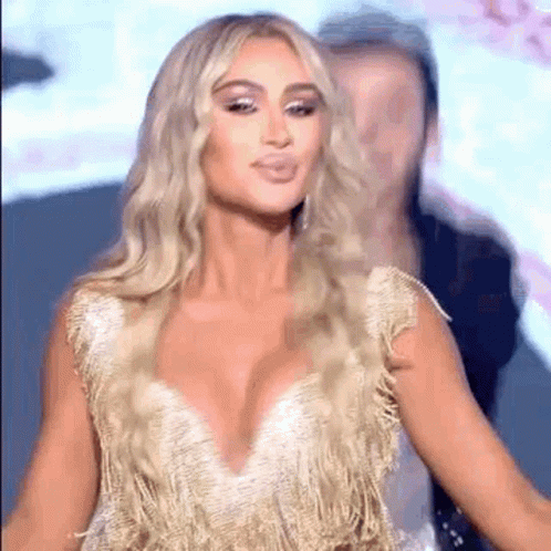 A Woman In A Gold Dress With A Plunging Neckline Is Standing In Front Of A Man . GIF