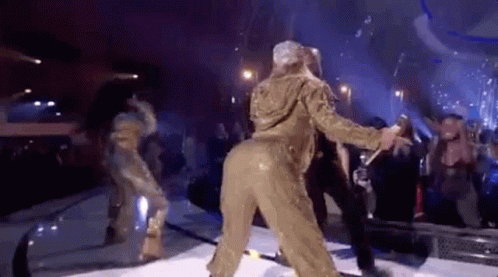 A Woman In A Gold Jumpsuit Is Dancing On A Stage In Front Of A Crowd . GIF