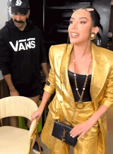 A Woman In A Gold Suit Is Standing Next To A Man Wearing A Vans Shirt GIF