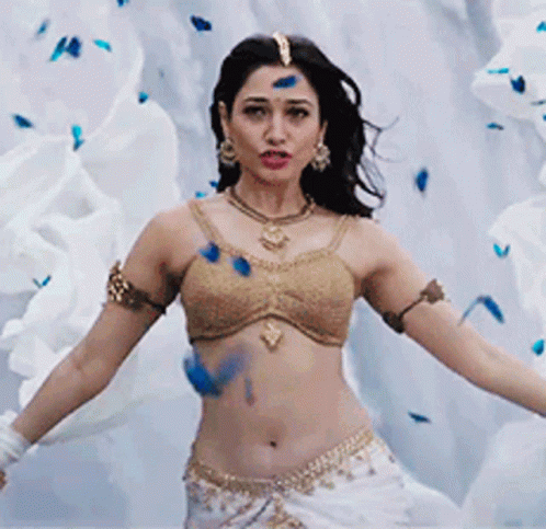 A Woman In A Gold Top And White Skirt Is Dancing In Front Of A White Wall . GIF