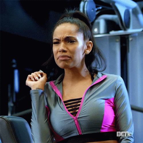 A Woman In A Gray And Pink Sports Bra Is Sitting In A Gym Making A Funny Face GIF