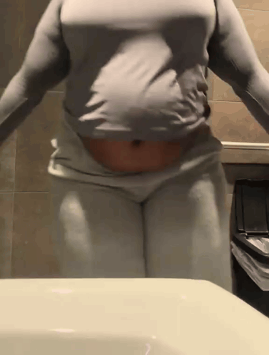 A Woman In A Gray Shirt And White Pants Is Standing In Front Of A Bathroom Sink . GIF