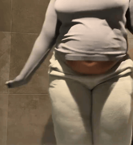 A Woman In A Gray Shirt And White Pants Is Standing In Front Of A Tiled Wall . GIF