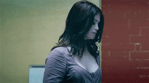 A Woman In A Gray Shirt Is Standing In Front Of A Red Wall . GIF