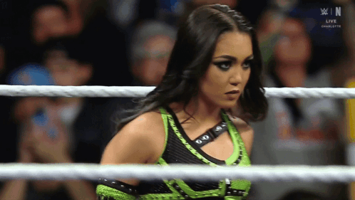 A Woman In A Green And Black Outfit Is In A Wrestling Ring With The Letters N On The Screen Behind Her GIF