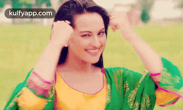 A Woman In A Green And Yellow Dress Is Smiling While Holding Her Hair . GIF