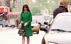 A Woman In A Green Coat And Sunglasses Is Standing On A Busy City Street . GIF