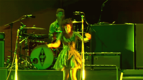 A Woman In A Green Dress Is Singing Into A Microphone In Front Of A Drum Set With The Letters Jbf On It GIF