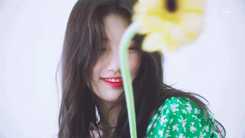 A Woman In A Green Dress Is Smiling While Holding A Yellow Flower In Front Of Her Face GIF