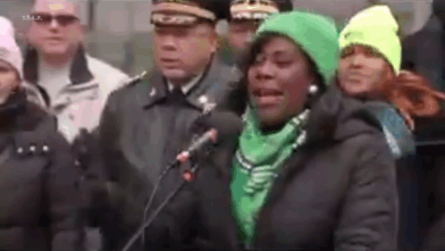 A Woman In A Green Hat Is Speaking Into A Microphone While Standing In Front Of A Crowd Of People . GIF