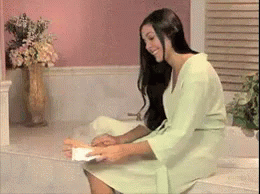A Woman In A Green Robe Is Sitting On A Toilet Holding A Piece Of Toilet Paper GIF
