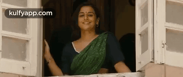 A Woman In A Green Sari Is Standing In A Window And Smiling . GIF
