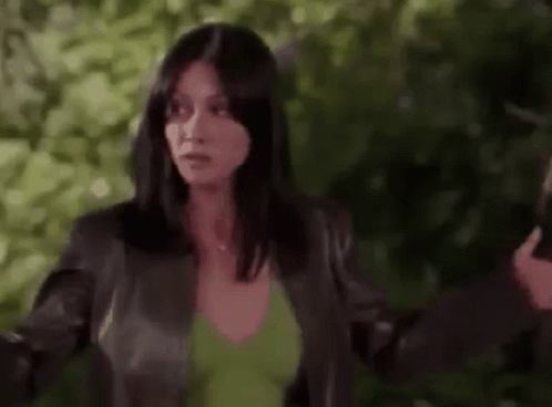 A Woman In A Green Shirt And A Black Jacket Is Standing In Front Of A Tree . GIF