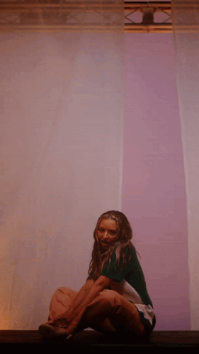 A Woman In A Green Shirt Sits On A Purple Wall GIF
