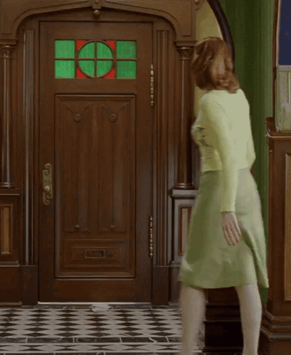 A Woman In A Green Skirt Is Walking In Front Of A Door With A Stained Glass Window GIF