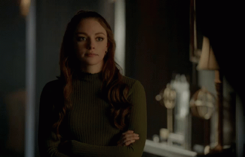 A Woman In A Green Sweater Stands With Her Arms Crossed In A Dark Room GIF