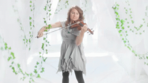 A Woman In A Grey Dress Is Playing A Violin In Front Of A White Curtain GIF