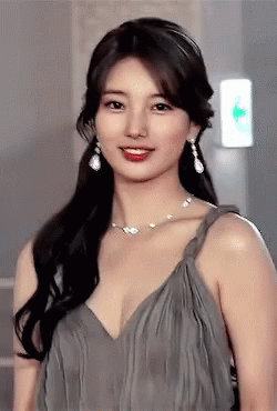 A Woman In A Grey Dress With A Necklace And Earrings Looks At The Camera GIF