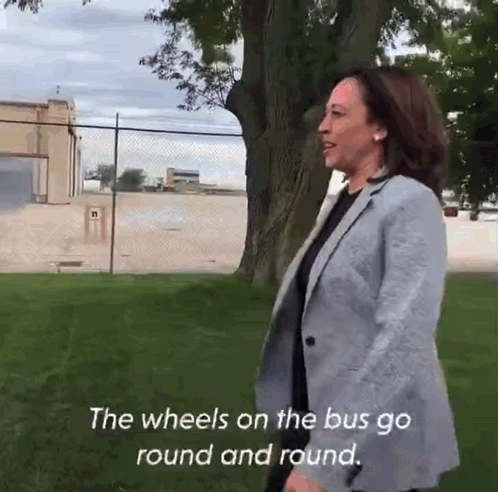 A Woman In A Grey Jacket Is Standing In A Grassy Field With The Words " The Wheels On The Bus Go Round And Round " Below GIF
