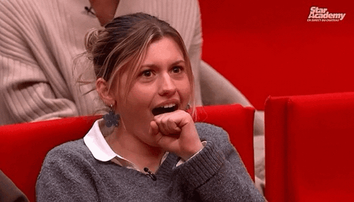 A Woman In A Grey Sweater Is Sitting In A Red Chair With A Star Academy Logo Behind Her GIF