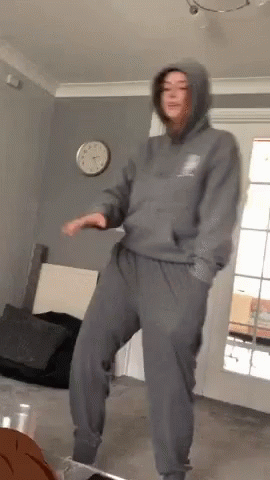 A Woman In A Hoodie And Sweatpants Is Dancing In A Living Room GIF