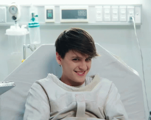 A Woman In A Hospital Gown Is Smiling And Looking At The Camera While Sitting In A Bed GIF