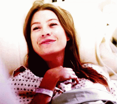 A Woman In A Hospital Gown Smiles While Laying In A Hospital Bed GIF