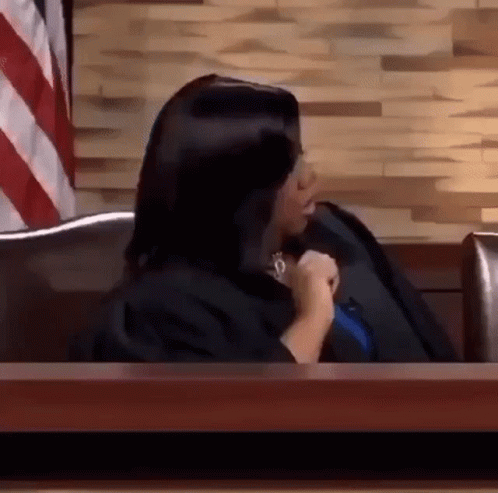 A Woman In A Judge 'S Robe Is Sitting At A Table In Front Of A Wooden Wall . GIF