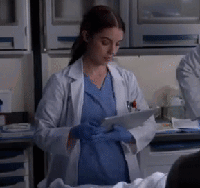 A Woman In A Lab Coat And Blue Scrubs Is Using A Tablet GIF