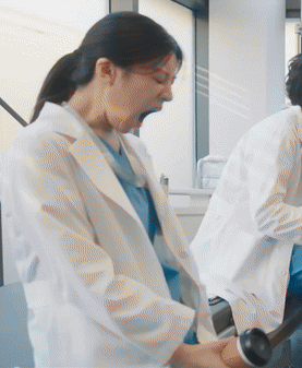 A Woman In A Lab Coat Is Yawning While A Man In A Lab Coat Is Using A Machine . GIF