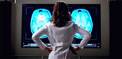A Woman In A Lab Coat Stands In Front Of A Large Screen Showing A Brain Scan GIF