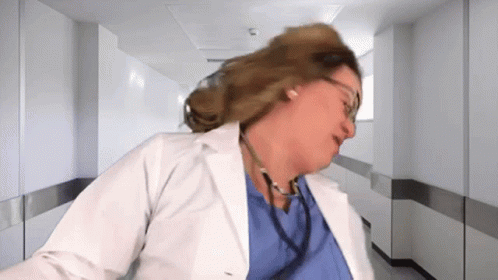A Woman In A Lab Coat With A Stethoscope Around Her Neck Is Standing In A Hospital Hallway . GIF