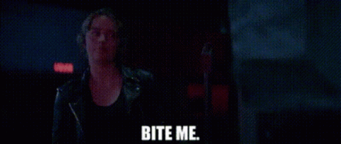 A Woman In A Leather Jacket Is Standing In A Dark Room With The Words Bite Me Written On The Screen . GIF