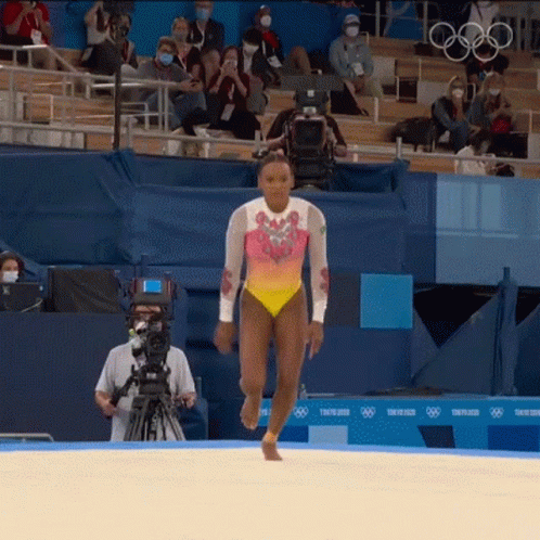 A Woman In A Leotard Is Walking On A Sandy Surface In Front Of A Tokyo 2020 Sign GIF
