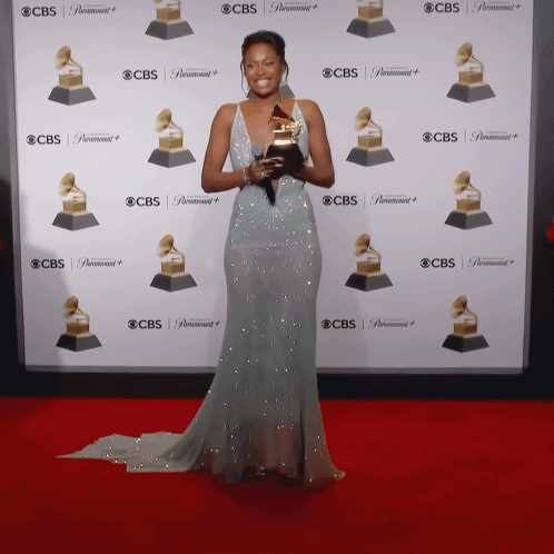 A Woman In A Long Dress Holds A Grammy Trophy In Front Of A Wall That Says Cbs GIF