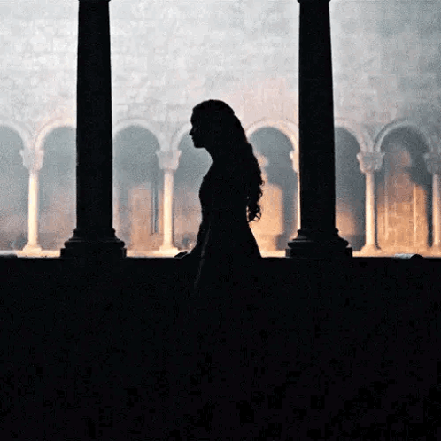 A Woman In A Long Dress Is Standing In Front Of A Window In A Dark Room . GIF