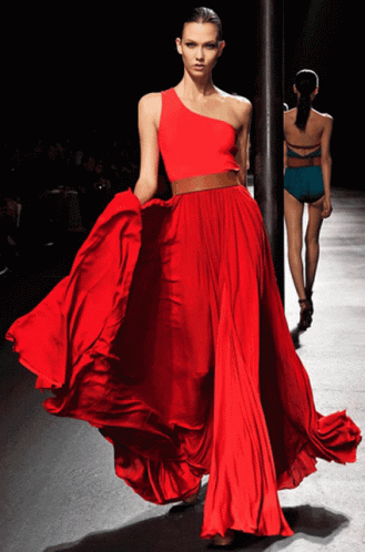 A Woman In A Long Red Dress Is Walking Down A Runway GIF