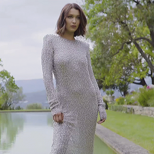 A Woman In A Long Silver Dress Is Standing In Front Of A Body Of Water GIF