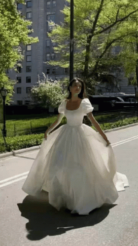A Woman In A Long White Dress Is Walking Down A Street GIF