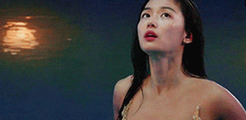 A Woman In A Mermaid Costume Is Standing In Front Of A Body Of Water Looking Up . GIF