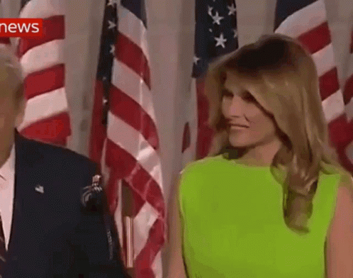A Woman In A Neon Green Dress Is Standing Next To A Man In A Suit In Front Of American Flags . GIF
