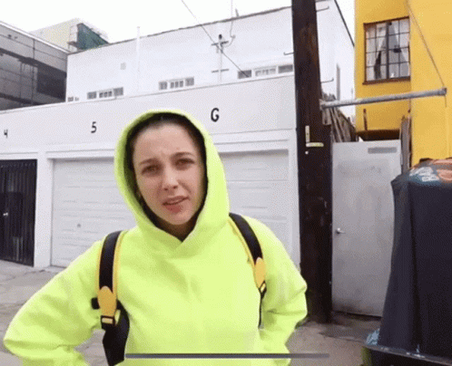 A Woman In A Neon Yellow Hoodie Stands In Front Of A Building With The Number G On It GIF