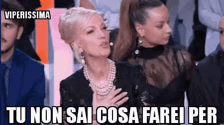A Woman In A Pearl Necklace Is Sitting In Front Of A Group Of People With Her Hand On Her Chest . GIF