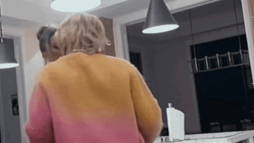 A Woman In A Pink And Orange Sweater Is Standing In A Kitchen Looking At Herself In The Mirror . GIF