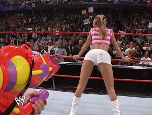 A Woman In A Pink And White Striped Top Is Dancing In A Wrestling Ring While A Man Holds A Water Gun GIF
