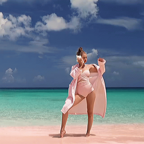 A Woman In A Pink Bathing Suit Is Standing On A Beach Holding A Seashell GIF