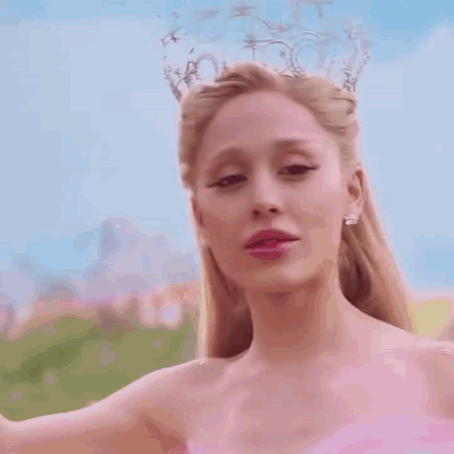 A Woman In A Pink Dress And A Crown On Her Head Is Standing In Front Of A Blue Sky . GIF