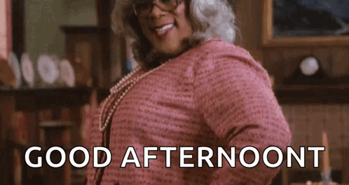 A Woman In A Pink Dress And Glasses Is Standing In A Room And Smiling . GIF
